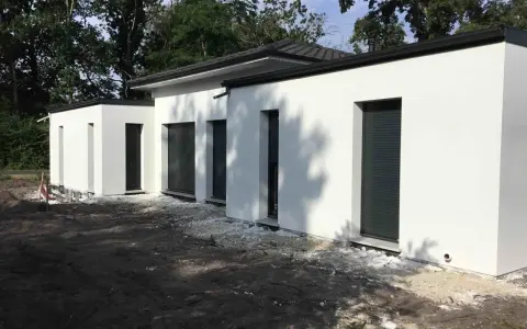 Maison moderne avec toit plat, plan de 150m2. Construction blanche aux lignes épurées, entourée d'arbres.
