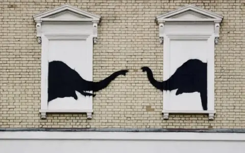 Œuvre Banksy : deux éléphants noirs se touchent la trompe à travers deux fenêtres sur un mur de briques.