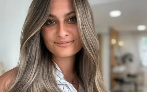 Frau mit langen, welligen Haaren in Balayage-Optik, aschblond mit helleren Strähnen. Ihr Blick ist warm und einladend.