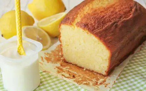 Bizcocho de yogur casero, dorado y esponjoso, acompañado de limones frescos y un vasito de yogur.