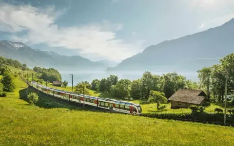 A train travels through a lush green valley, with a lake and mountains in the background. This scenic route from Lucerne to Montreux offers breathtaking views.
