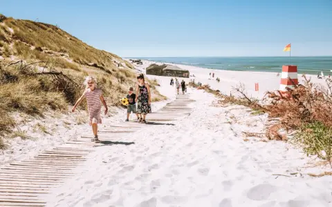 Familj promenerar på en strand med vit sand i Danmark. En livräddartorn och bunkrar syns i bakgrunden.