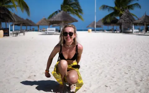 Uśmiechnięta blondynka w bikini i żółtej spódniczce kuca na piaszczystej plaży na Zanzibarze. W tle palmy i słomiane parasole.