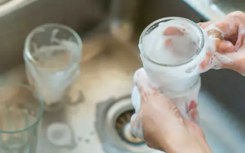 Händer tvättar glas med skum i diskho för att glaspest ta bort.