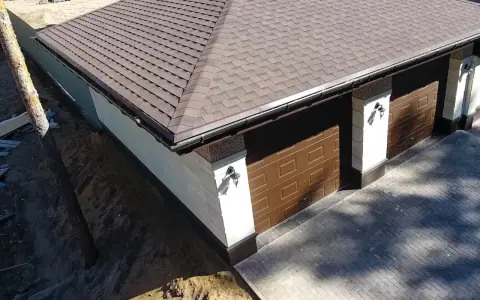 Nowy garaż z brązowym pokryciem dachowym bitumicznym. Eleganckie drzwi garażowe i ozdobne słupy.