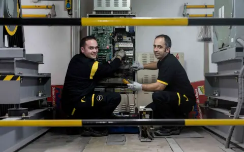 Técnicos realizan mantenimiento de elevadores en un taller, revisando la electrónica y cableado.