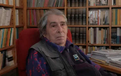 Ferdinando Camon, con i suoi capelli grigi e lo sguardo pensieroso, siede tra scaffali pieni di libri, un ritratto di un intellettuale immerso nel suo mondo.