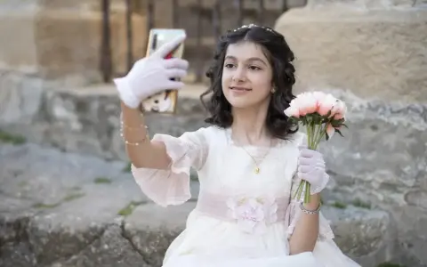 Dziewczynka w sukni komunijnej robi selfie z bukietem kwiatów. To ważny moment, przygotowania do pierwszej komunii świętej krok po kroku.