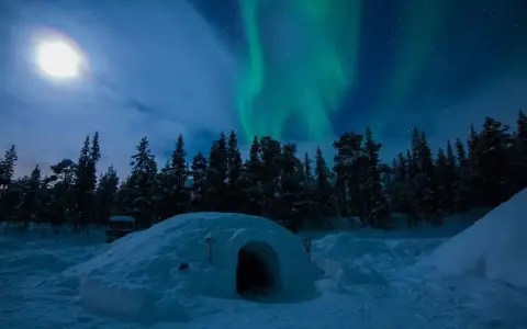 En glasigloo i Sverige under norrsken. En snöig skog och en fullmåne lyser upp natten.