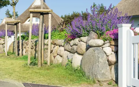 Natürliche Steingarten Ideen: Eine Steinmauer mit Lavendel und rosa Blüten, flankiert von jungen Bäumen und einem Reetdachhaus.