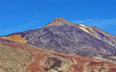 Majestätischer Vulkan im Teide Nationalpark unter blauem Himmel. Die Hänge zeigen verschiedene Gesteinsschichten und eine sandfarbene Fläche.