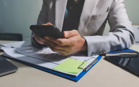 Osoba w jasnej marynarce przegląda telefon, na biurku dokumenty z wykresem i notatkę z informacją o uchyleniu zajęcia.