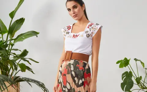 Mujer con top blanco bordado y falda plisada de estampado abstracto. Un cinturón marrón ancho realza la cintura.