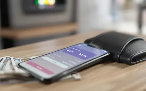 Smartfon z aplikacją BLIK, obok portfel i banknoty na drewnianym stole.