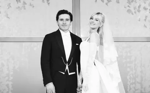 Novios sonrientes en la boda Brooklyn Beckham. Él con esmoquin, ella con velo y vestido blanco.