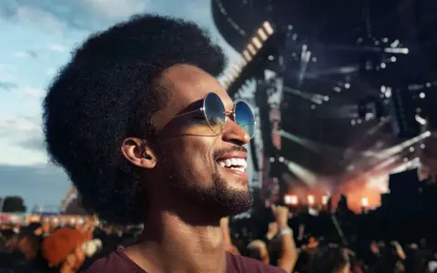 Homme souriant à un concert, portant des lunettes de soleil et un bouchon d'oreille pour protéger son audition.