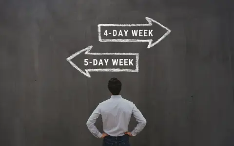 Mężczyzna stoi przed tablicą, na której kredą narysowano dwie strzałki: w prawo "4-DAY WEEK" i w lewo "5-DAY WEEK".