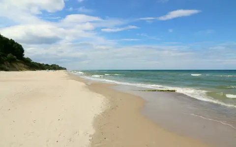 Plaża nad morzem, gdzie fale delikatnie obmywają piasek. W oddali widać zalesione wzgórze, a na niebie chmury. Idealne miejsce na spacer dla gąsek.