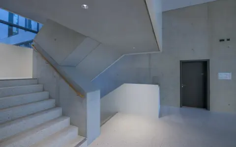 Moderne Treppe mit Sichtbeton-Wänden, die durch die Schalung eine interessante Textur erhalten. Ein dunkler Türrahmen und ein Holzhandschuh ergänzen das Bild.