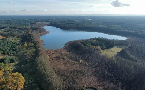 Malownicze jezioro Wolickie otoczone lasami i polami. Spokojna tafla wody odbija błękitne niebo z chmurami.