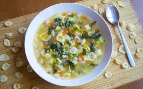 Un reconfortante plato de sopa minestrone, lleno de pasta orecchiette, verduras y frijoles, servido en un tazón blanco sobre una tabla de madera.