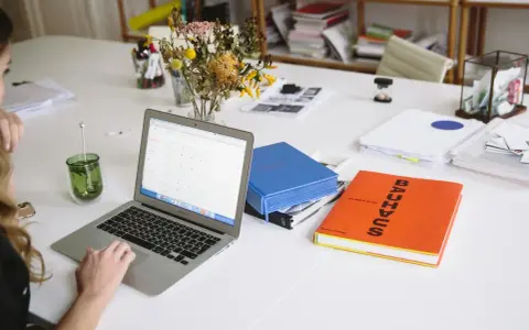 Escritorio con portátil, libros y flores. Un espacio de trabajo que parece una copistería por la cantidad de documentos y la organización.