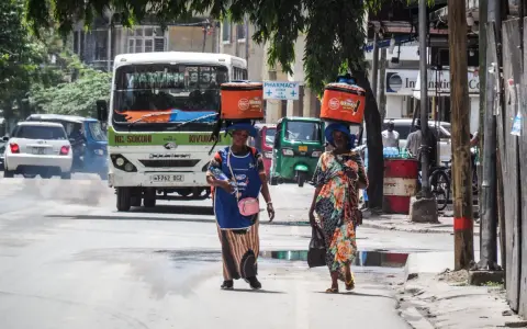 Dwie kobiety niosą na głowach pomarańczowe lodówki w upalny dzień w Dar es Salaam. W tle autobus i ruchliwa ulica.
