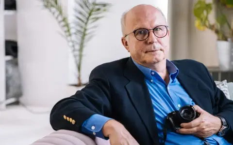 Tomaszewski fotograf w okularach, w granatowej marynarce i niebieskiej koszuli, trzyma aparat.