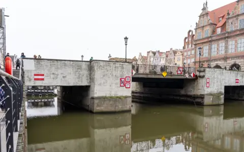 Nad kanałem w Gdańsku wznosi się betonowy zielony most, ozdobiony czerwono-białymi znakami. Ludzie spacerują po jego balustradzie.