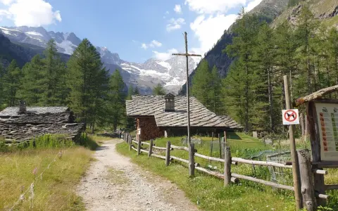 Sentiero di montagna con baite in pietra e tettoie, ideale per cogne escursioni facili. Montagne innevate sullo sfondo.