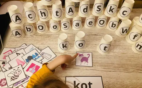 Dziecko układa wieżę z kubeczków z literami, ucząc się jak nauczyc dziecko liter. Obok leżą obrazki, w tym kot, który pasuje do słowa "kot".