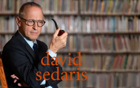 David Sedaris, con pipa in mano, seduto tra scaffali pieni di libri, riflette pensieroso.