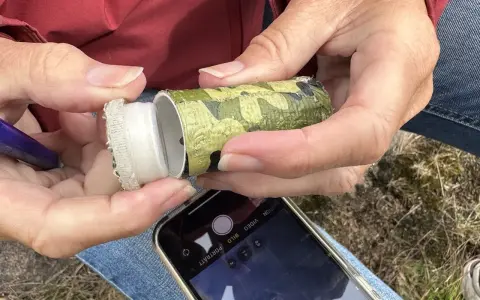 Händer öppnar en kamouflerad behållare, redo för geocaching i Sverige. En mobiltelefon ligger redo att dokumentera fyndet.