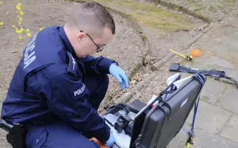 Policjant w niebieskim mundurze z napisem "POLICJA" klęczy przy otwartej walizce z aparatem i sprzętem kryminalistycznym.