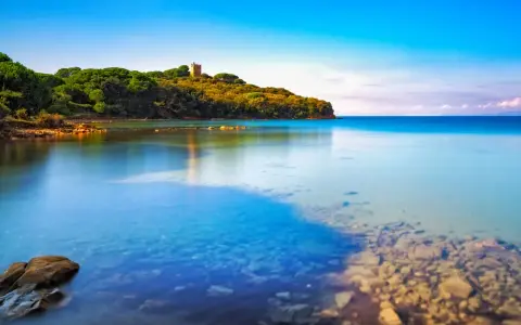 Torre medievale su una collina verdeggiante affacciata sul limpido **golfo di Follonica**. Acque calme e trasparenti riflettono il cielo azzurro.