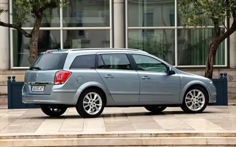Srebrny Opel Astra H 1.6 benzyna kombi zaparkowany na tle nowoczesnego budynku z drzewami.