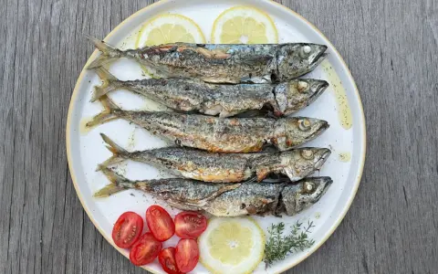 Cinq chinchards poissons grillés, servis sur une assiette avec des quartiers de citron, des tomates cerises et des herbes.