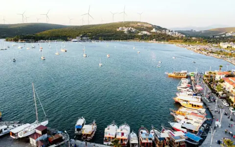 Port w Alacati, Turcja, pełen łodzi i żaglówek, z wiatrakami na wzgórzach w tle.
