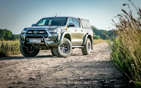 Złota toyota terenowa z wyciągarką i zabudową pędzi po polnej drodze, otoczona dziką roślinnością.