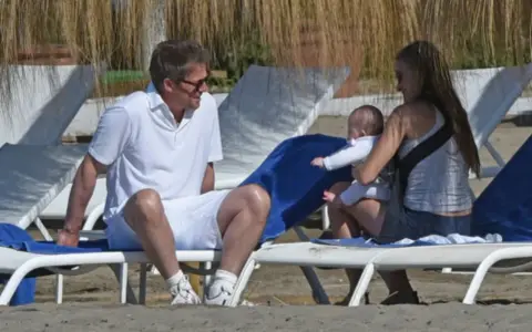 Hugh Grant njuter av stranden med sitt barn och en kvinna.