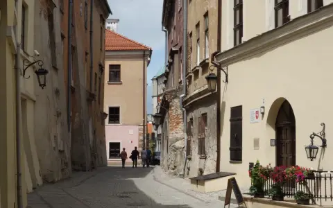 Wąska, brukowana uliczka w Lublinie z kamienicami i kilkoma spacerującymi osobami.