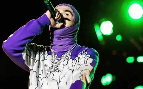 Raper w fioletowej bluzie z kapturem i naszyjnikiem z krzyżem śpiewa do mikrofonu. Zielone światła sceniczne tworzą atmosferę. Mata Tango tekst.