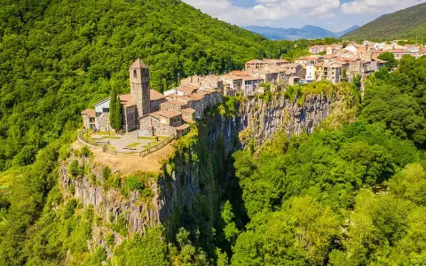 Un borgo medievale, Castellfollit de la Roca, aggrappato a una scogliera basaltica, circondato da lussureggianti foreste verdi.