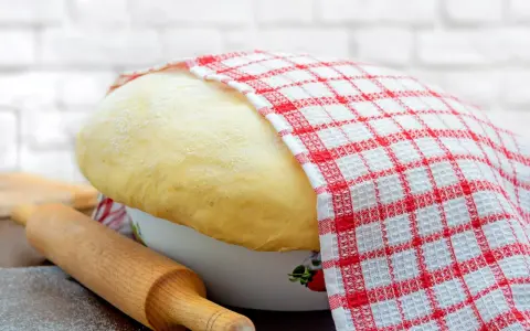 Wyrośnięte ciasto drożdżowe, przykryte ręcznikiem w kratkę, czeka na swój przepis babci. Obok wałek i mąka.