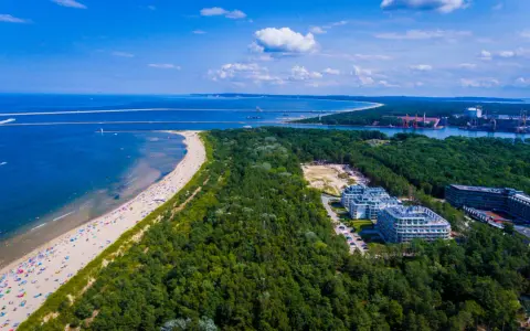 Widok z lotu ptaka na najszerszą plażę w Polsce, pełną wypoczywających ludzi, otoczoną lasem i nowoczesnymi budynkami.