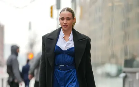 Frau trägt eine schwarze Jacke über einem blauen Nadelstreifen-Jumpsuit mit Korsett-Oberteil und weißem Hemd.