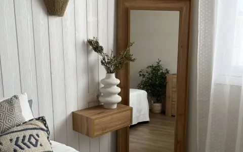 Estantería flotante de madera con jarrón blanco y ramas secas, ideal para ideas para decorar rincones pequeños. Reflejo de planta y cama en espejo grande.