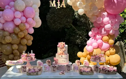 Mesa dulce baby shower con pastel de osito, galletas temáticas y globos rosas y dorados. ¡Bienvenida Mia!