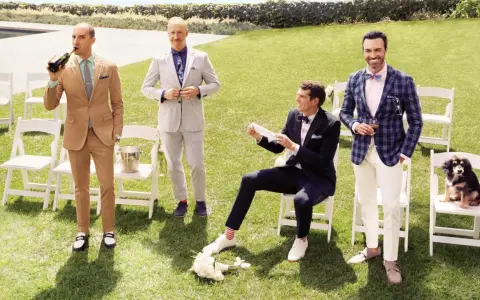 Four men in stylish garden formal wedding attire for male guests pose on a lawn. One drinks champagne, another adjusts his suit, one sits with a bouquet, and the last holds a drink with a dog nearby.