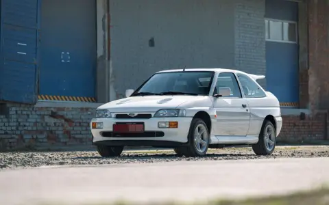 Biały Ford Escort Cosworth z charakterystycznym spojlerem, zaparkowany przed industrialnym budynkiem z niebieskimi drzwiami.